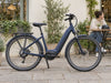 Navy step-through e-bike parked beside a leafy cafe terrace with its rider in the background. AI-generated example.