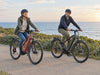 Two helmeted adults riding unbranded e-bikes along a coastal path beside the sea. AI-generated lifestyle example.
