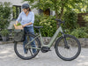 Helmeted woman placing groceries into the rear pannier of a sage-green step-through e-bike. AI-generated example.