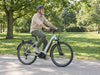 Older adult wearing a helmet riding a silver step-through e-bike along a park path. AI-generated lifestyle example.