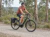 Helmeted rider on a green trekking e-bike with a rear pannier on a gravel woodland trail. AI-generated example.