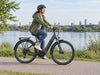 Woman wearing a helmet riding a charcoal step-through e-bike beside a river. AI-generated lifestyle example.